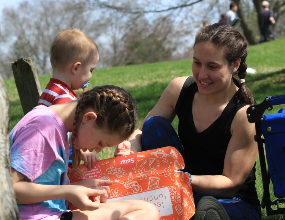 Christina Grubb, of Newark, sitting with Wyatt Grubb, 2, and Tori Reifsnyder, 10, opens the learning lunchbox distributed as a part of events surrounding the solar eclipse at The Dawes Arboretum on Monday, April 8, 2024.