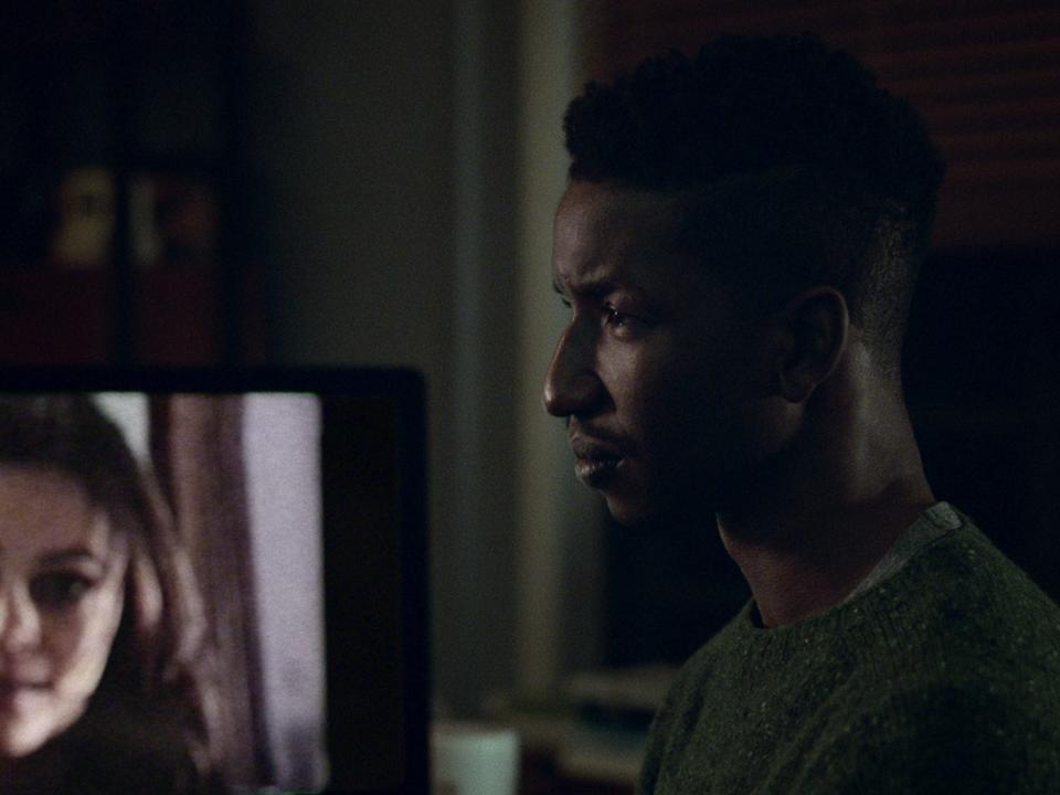 Mamoudou Athie standing in front of screen with woman's face on it in "Archive 81"