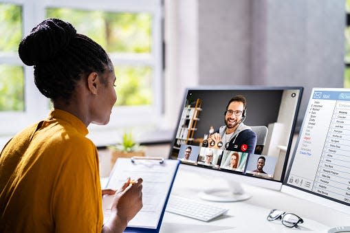 A woman in a yellow shirt sitting at a desk and writing down notes on a clipboard while listening during a virtual meeting.