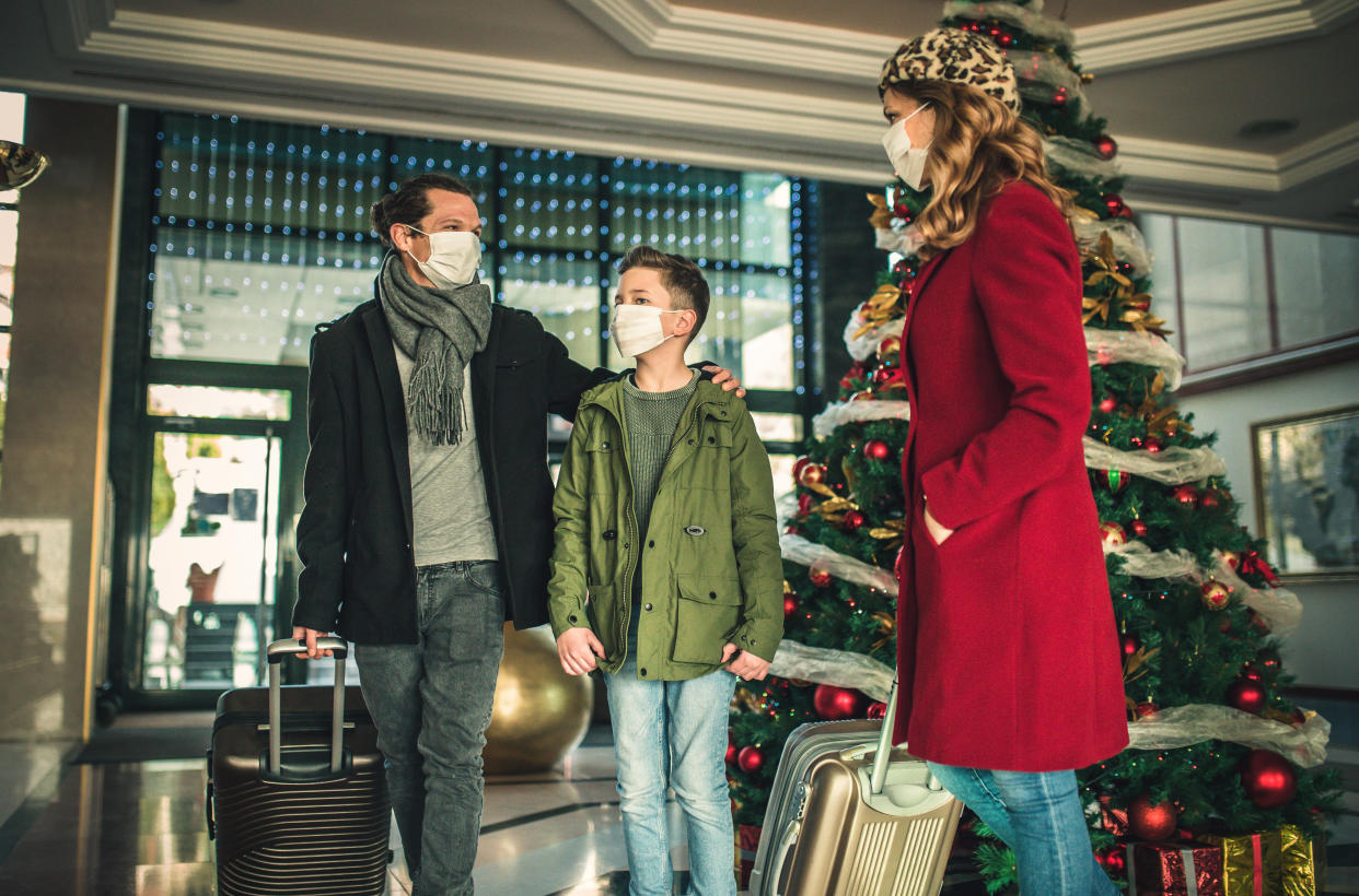 A family of parents and their young son, with suitcases, all wearing masks and outdoor clothes, stand in a commercial area decorated with a Christmas tree.