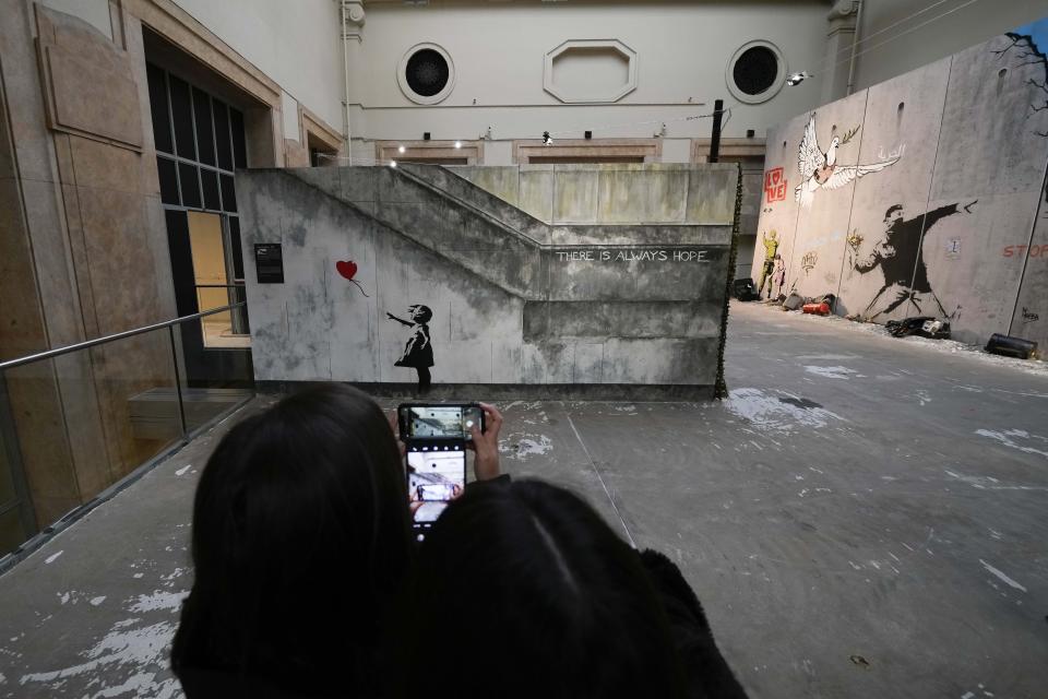 Visitors take pictures to "Girl with balloon", a reproduction of a mural by British artist Banksy, during the unveiling of the "The World of Banksy, The Immersive Experience" exhibition, in Milan, Italy, Thursday, Dec. 2, 2021. An exhibition of 130 works by British street artist Banksy opens Friday in a gallery space inside Milan's Central train Station. The exhibition unveiled on Thursday includes 30 never before seen works by Bansky and highlights pieces by young unknown artists from all over Europe. (AP Photo/Luca Bruno)