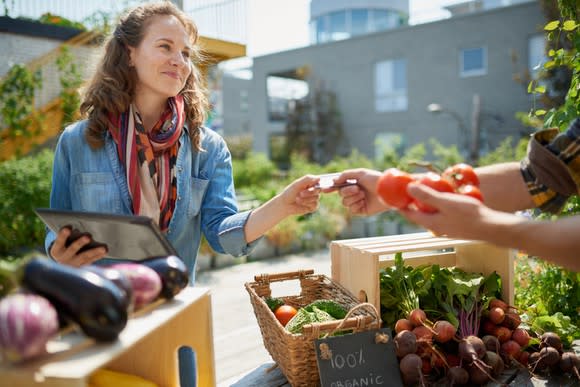 Smiling female merchant selling goods at a farmers market, using a mobile checkout device