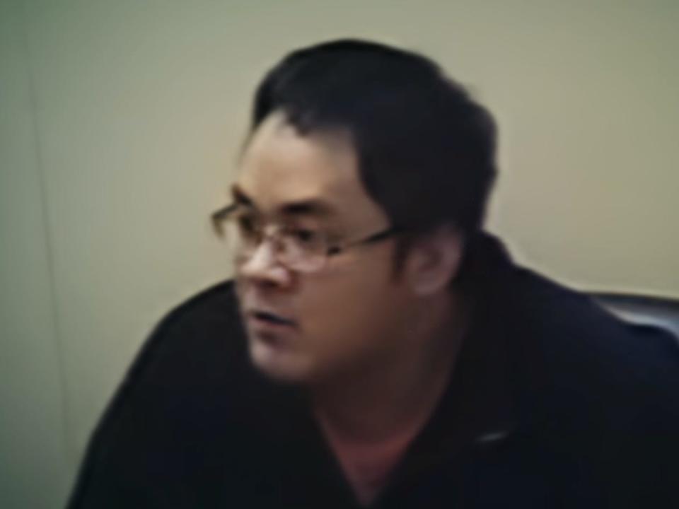 daniel wong in police interview footage, wearing a balck shirt and glasses, sitting in a chair. the image quality is poor