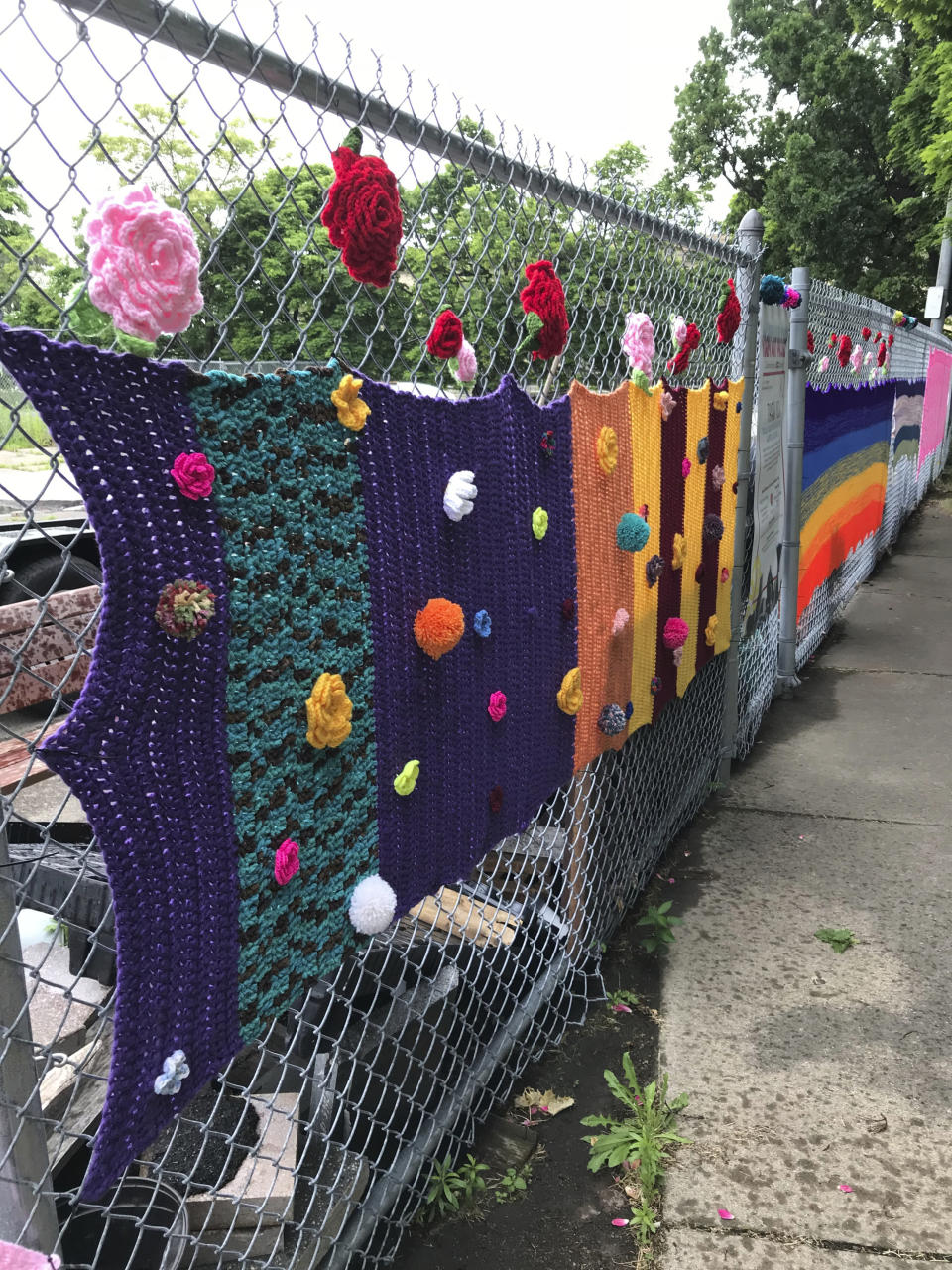 This June 2018 photo provided by Hinda Mandell shows a yarn installation at Schiller Park in Rochester, N.Y. More than 40 women from Rochester participated in "Yarn at Schiller Park," a community event organized by Mandell to spruce up Schiller Park. Dozens of knit and crocheted blankets decorated chain-link fencing to bring color to a neglected park that bordered an outdoor storage space for the city's construction equipment. Knitters and crocheters call it yarn bombing. They're using fiber arts to make political statements, or maybe just to lift people's spirits. Experts say yarn bombing is part of a long tradition in which women use textile arts to agitate, excite or inspire. (Hinda Mandell via AP)