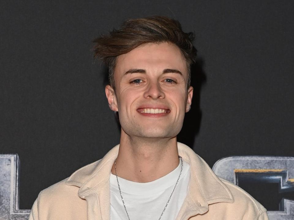 Joe Tasker smiling while wearing a white shirt and cream-colored jacket and posing in front of a black background.