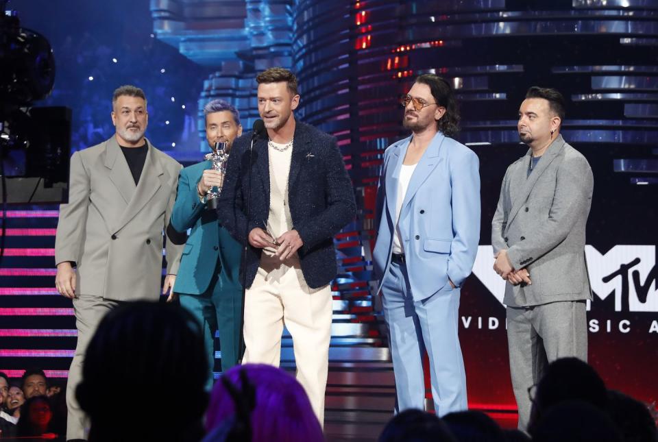 joey fatone, lance bass, justin timberlake, jc chasez, and chris kirkpatrick stand on a stage as timberlake speaks into a microphone, all five men wear suits