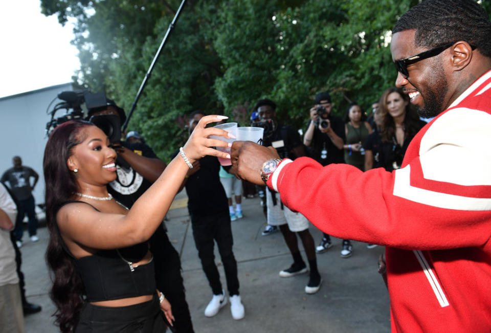 I don't know who these people are. Two people, one in a black outfit and the other in a red and white jacket, exchanging drinks with a group of photographers in the background