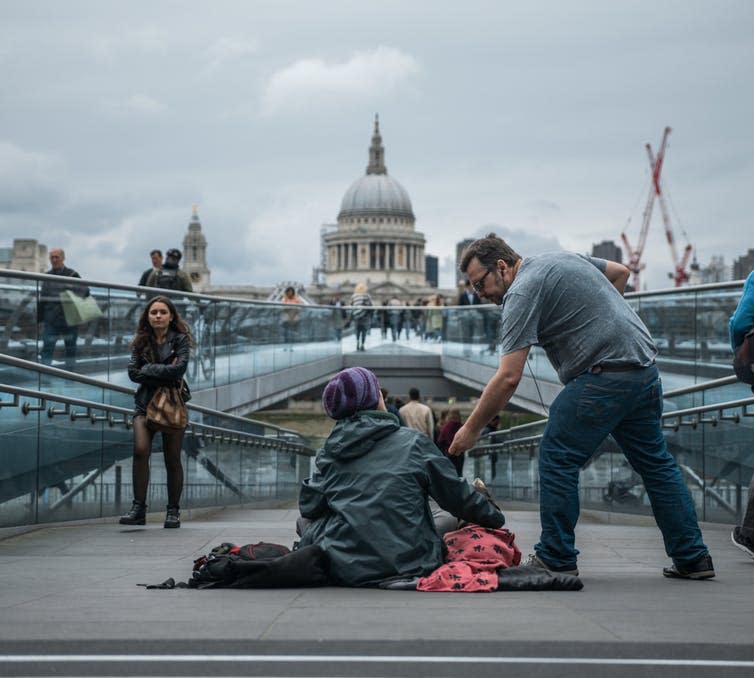 A man in a grey t-shirt gives money to a homeless person sitting on a bridge in central London