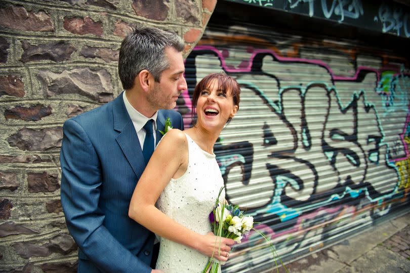 Bride and groom embrace while stood in front of a shutters with graffiti