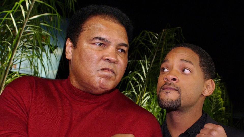 muhammad ali and will smith pose for a photo with each holding one fist up on their chest, ali wears a red long sleeved shirt and smith wears a black shirt