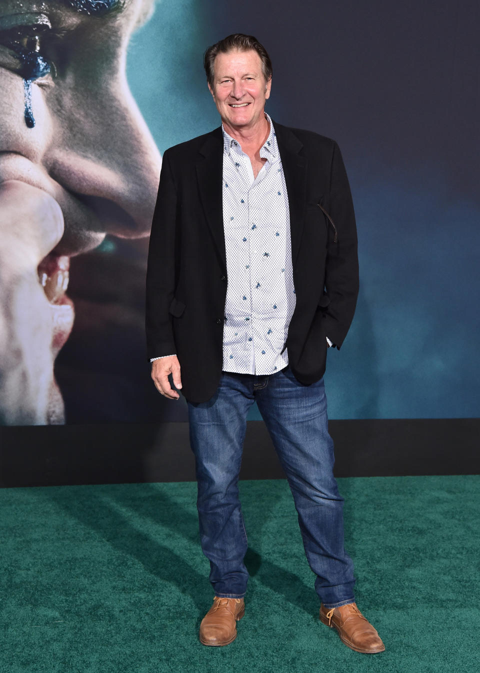 Brett smiles at the Joker premiere, wearing a black blazer jacket over a white shirt with small navy details, dark blue jeans and tan colour shoes