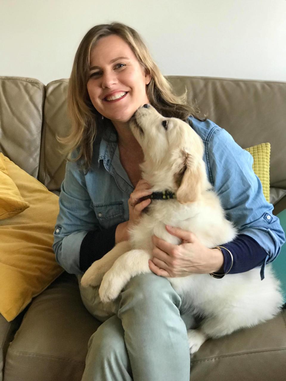 Dr Leigh Davidson sits on a couch holding a dog that is licking her chin. 