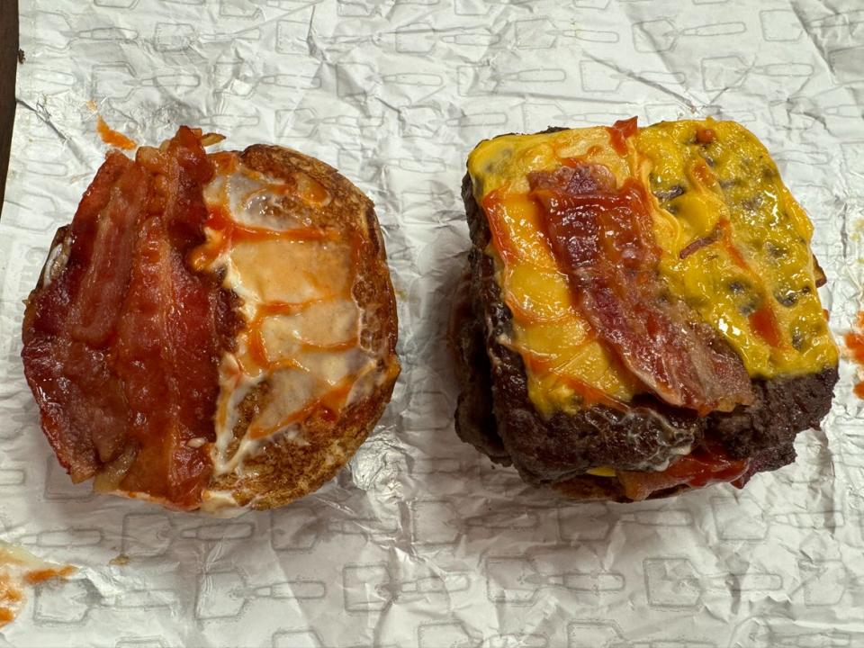 A burger with its bun taken off and placed on the side of it. The bun has a piece of bacon and sauce and the side with the burger is covered with cheese, sauce, and a piece of bacon