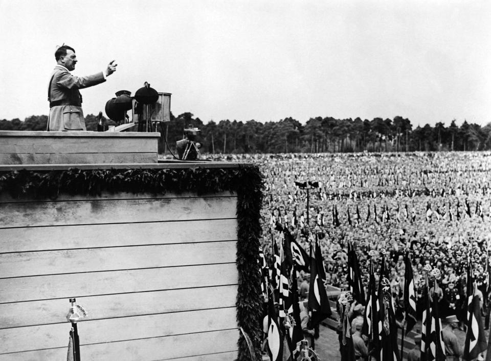 Adolf Hitler delivers a speech to a large, uniformed crowd, raising his arm in a salute, during a rally