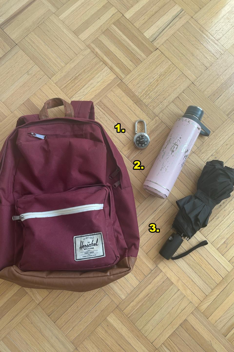 A backpack, a combination lock, a water bottle, and an umbrella are placed on a wooden floor. The backpack has a label saying 