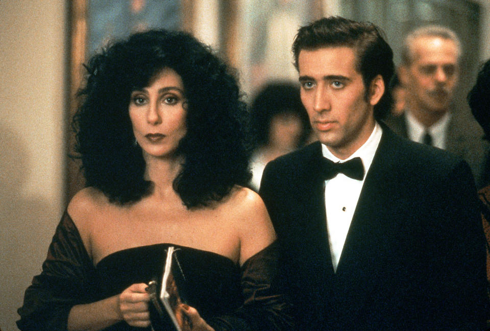 Cher and Nicolas Cage, dressed in formal attire, are walking together at an event. Cher wears an off-shoulder gown, and Cage is in a black tuxedo