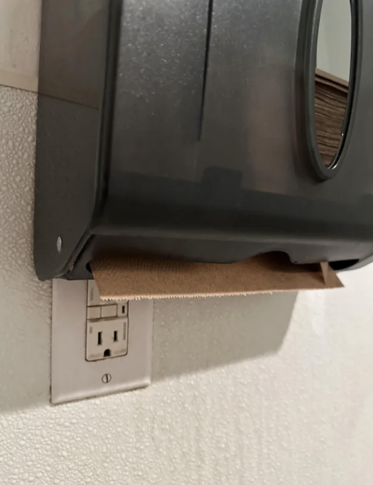 An electrical outlet is directly below a paper towel dispenser in a public bathroom