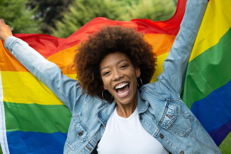 Cinematic shot of happy young african woman carrying flag of LGBT rainbow symbol and smiling in camera in green city park. Concept of homophobia, diversity, equity, peace and love, freedom, liberty.