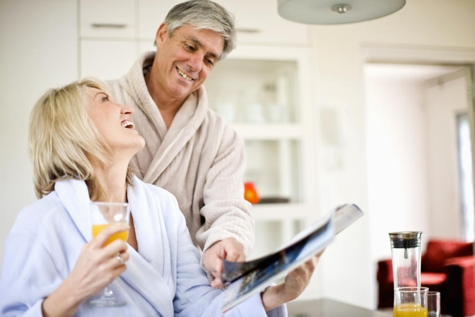 Senior man and woman in bathrobes; woman is holding glass of juice in one hand and magazine in other, while man points to magazine