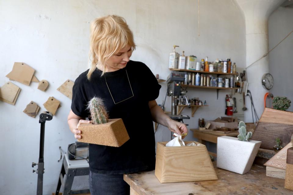 A woman holds a wood planter with a cactus in her right hand and touches a planter on a table with her left.