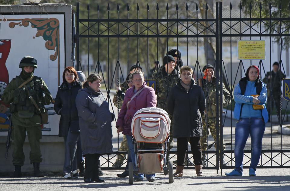 A military personnel member (L), believed to be a Russian serviceman, and women gather outside a Ukrainian military unit in the village of Perevalnoye, as Ukrainian servicemen are seen behind the gate, outside Simferopol March 3, 2014. Ukraine mobilised for war on Sunday and Washington threatened to isolate Russia economically after President Vladimir Putin declared he had the right to invade his neighbour in Moscow's biggest confrontation with the West since the Cold War. Russian forces have already bloodlessly seized Crimea, an isolated Black Sea peninsula where Moscow has a naval base. REUTERS/David Mdzinarishvili (UKRAINE - Tags: POLITICS MILITARY CIVIL UNREST)