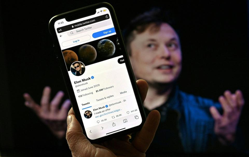 In this photo illustration, a phone screen displays the Twitter account of Elon Musk with a photo of him shown in the background, on April 14, 2022, in Washington, DC. - Tesla chief Elon Musk has launched a hostile takeover bid for Twitter, insisting it was a 