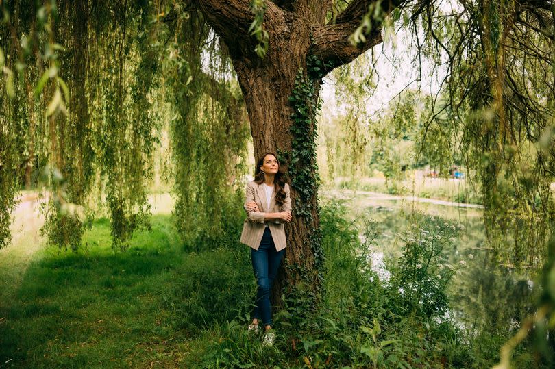 Kate Middleton stands under a tree outdoors, wearing a beige blazer over jeans