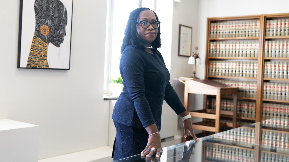 Judge Ketanji Brown Jackson stands in her office with law volumes in bookshelves to her left, and an artist's image of a Black woman on the wall behind her.