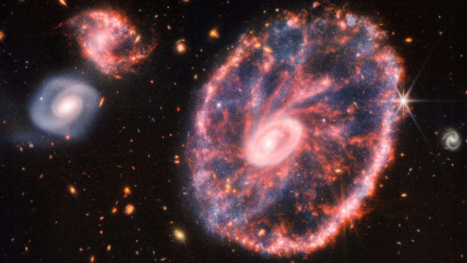 A photograph of the Cartwheel Galaxy shows it as pink swirls around a center, with other galaxies of all sizes around it, taken by the James Webb Space Telescope