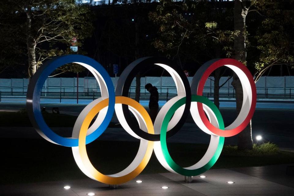 A man is seen through the Olympic rings in front of the New National Stadium in Tokyo, Tuesday, March 24, 2020. IOC President Thomas Bach has agreed "100%" to a proposal of postponing the Tokyo Olympics for about one year until 2021 because of the coronavirus outbreak, Japanese Prime Minister Shinzo Abe said Tuesday.
