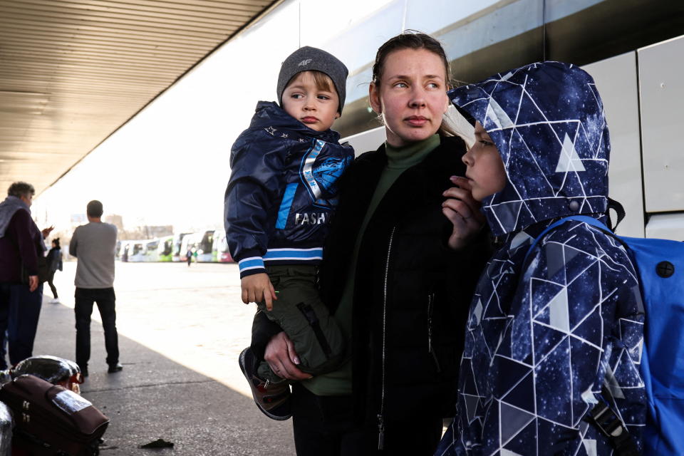 A young mother carrying a toddler waits in a bus shelter with her other son by her side.