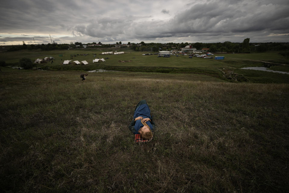 A woman taking part in the Romula Fest historic reenactment festival rests on a hillside, backdropped by replicas of Dacian and Roman camps, set up in the village of Resca, Romania, Saturday, Sept. 3, 2022. (AP Photo/Andreea Alexandru)