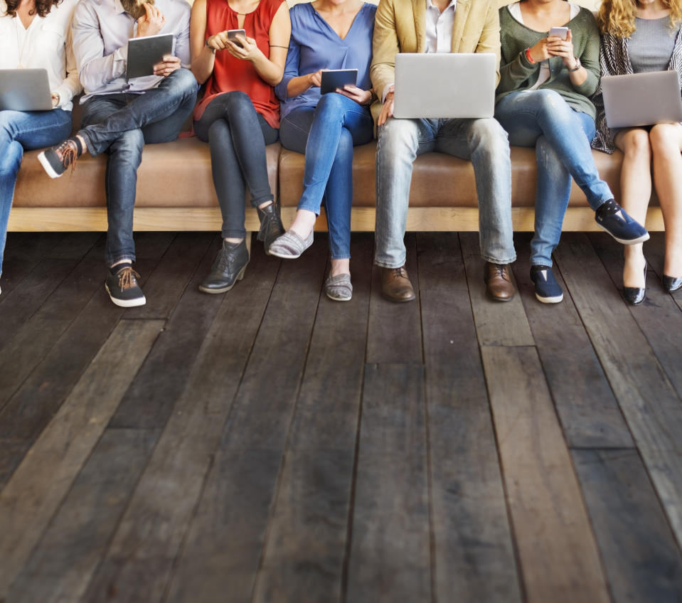 A group of people sitting on a bench working on laptops, tablets, and smartphones.