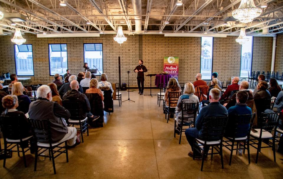 A small crowd, seen from behind, sits in rows in a large room as a man in black stands facing them, gesturing as he speaks.