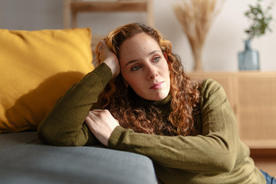 A woman resting head on hand, looking thoughtful, lying on couch with yellow pillow, deep in thought