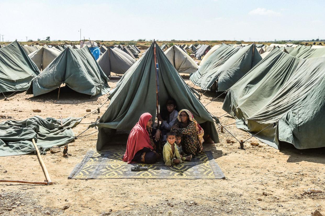 Two women with shawls over their heads and two children sit in the shelter of a tent on a mat set on parched ground.