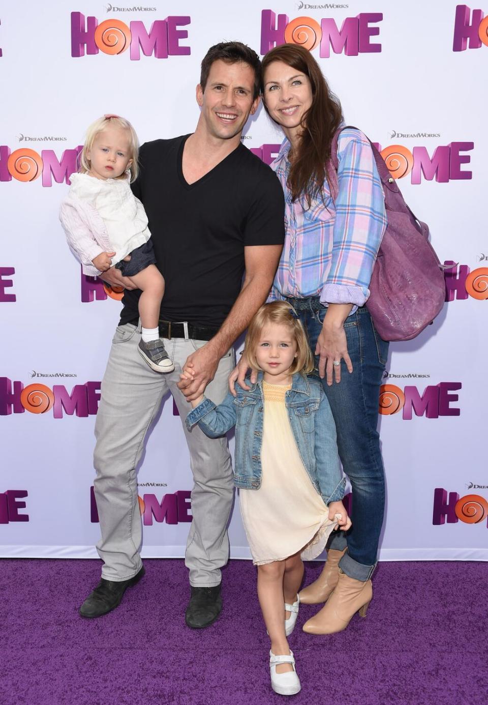 A man and a woman with long brown hair in casual clothing posing with two young children on a purple carpet
