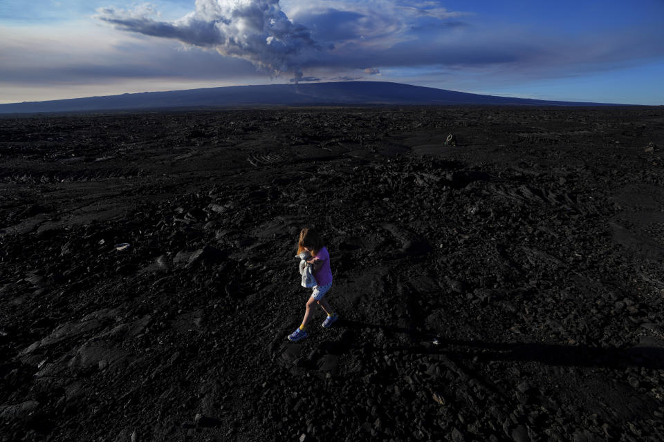 Abigail Dewar, of Alberta, Canada, holds a stuffed animal as she walks over hardened lava rock from a previous eruption as the Mauna Loa volcano erupts, behind, Wednesday, Nov. 30, 2022, near Hilo, Hawaii. (AP Photo/Gregory Bull)