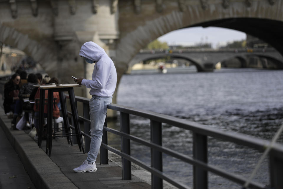 A man check his phone at a cafe terrace along the Seine river, Saturday Sept.26, 2020 in Paris. While France suffered testing shortages early in the pandemic, ramped-up testing since this summer has helped authorities track a rising tide of infections across the country. More than 15,000 new cases were reported Friday, and the Paris hospital system is starting to delay some non-virus surgeries to free up space for COVID-19 patients. (AP Photo/Lewis Joly)