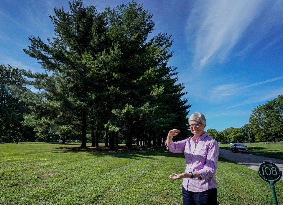 Marty Davis, who recently retired from Crown Hill Funeral Home and Cemetery after 40 years, gestures to show how tall the pine trees shown beside her were when they were planted around the time she began her career at the cemetery on Friday, August, 12, 2022, in Indianapolis.