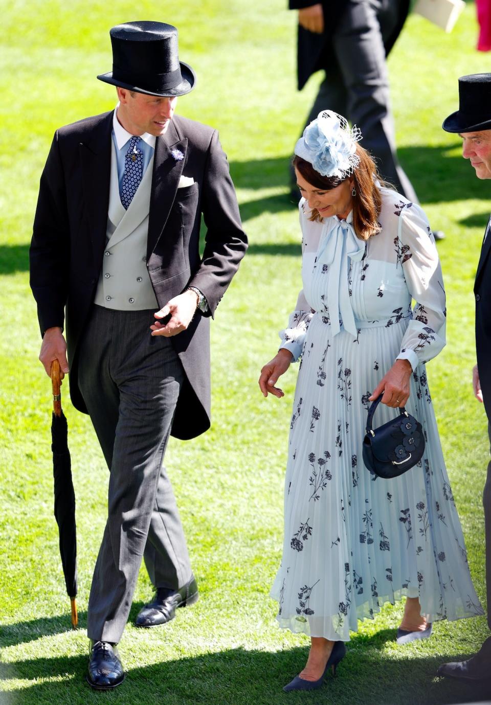 Prince William, wearing a black morning suit and top hat, walking with Carole Middleton, wearing a powder blue pleated dress with floral detailing.