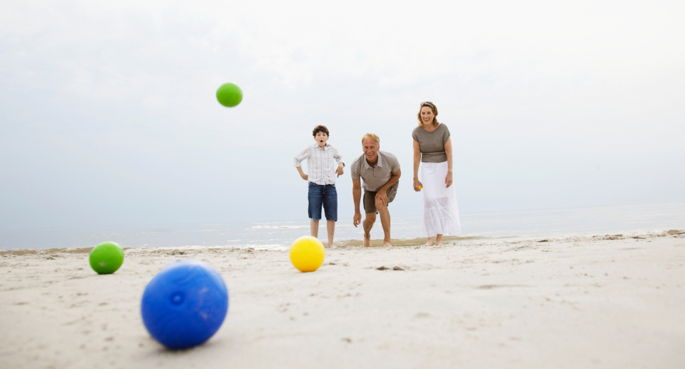 couple and child playing bocce ball with green balls, yellow ball and blue ball on sand at beach, outdoor games