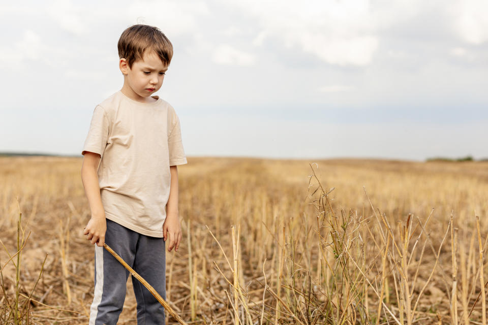 Young boy standing alone in an empty field, holding a stick and looking down pensively