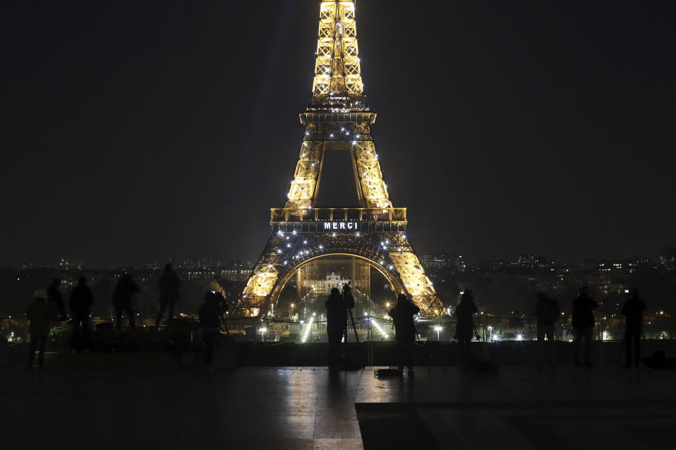 Media stand in front of the Eiffel Tower where the word "Merci", the French word for 'Thank you", is emblazoned as France's coronavirus death toll continued to climb, in Paris, Friday, March 27, 2020. Health workers fighting to save lives in France from COVID-19 have received a huge show of gratitude, from the Eiffel Tower. The new coronavirus causes mild or moderate symptoms for most people, but for some, especially older adults and people with existing health problems, it can cause more severe illness or death. (AP Photo/Thibault Camus)