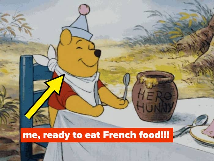 Winnie the Pooh sits at a table with a "HERO HUNNY" pot, wearing a bib and a party hat, happily holding a spoon, ready to eat with the caption: "me, ready to eat French food"