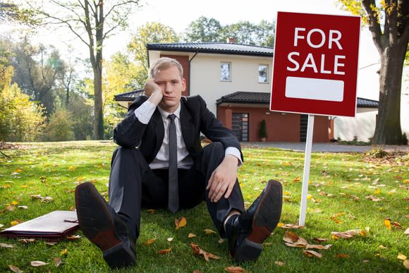 Unhappy man in suit sitting on lawn in front of a house and next to a for sale sign.