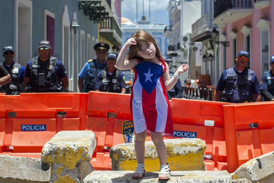 A girl wearing a dress featuring the Puerto Rican flag stands by police blocking the road leading to the La Fortaleza governors mansion in San Juan, Puerto Rico, Thursday, July 18, 2019. Protesters are demanding Gov. Ricardo Rossello resign after the leak of online chats that show him making misogynistic slurs and mocking his constituents. (AP Photo/Dennis M. Rivera Pichardo)