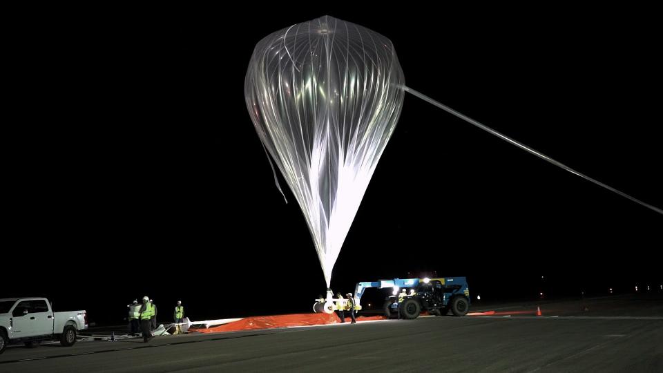 A crew at night hoists a large, clear balloon
