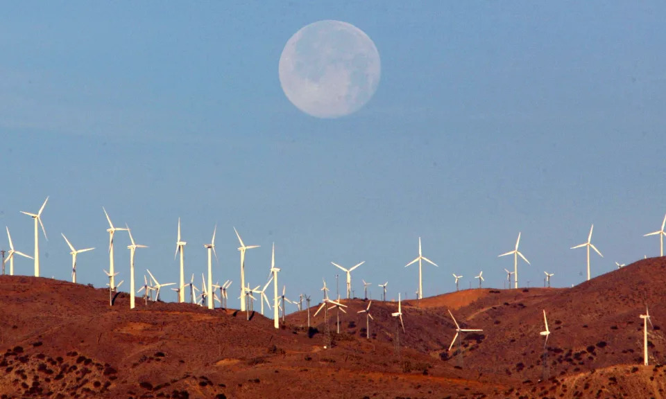 A full moon hovers near the horizon against a blue sky behind a wind farm with several dozen windmills visible in a mountainous area.