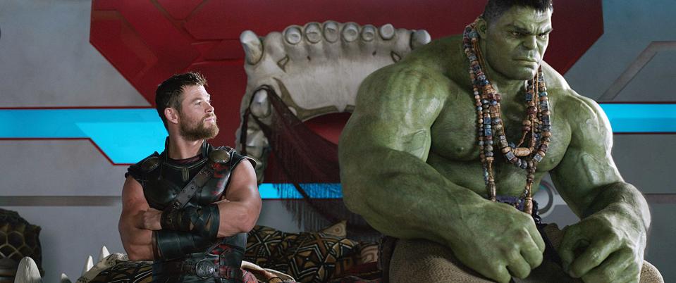 Chris Hemsworth as Thor and Mark Ruffalo as Hulk in a scene from a movie. Thor is in battle armor and Hulk, green and muscular, is wearing beads and sitting nearby